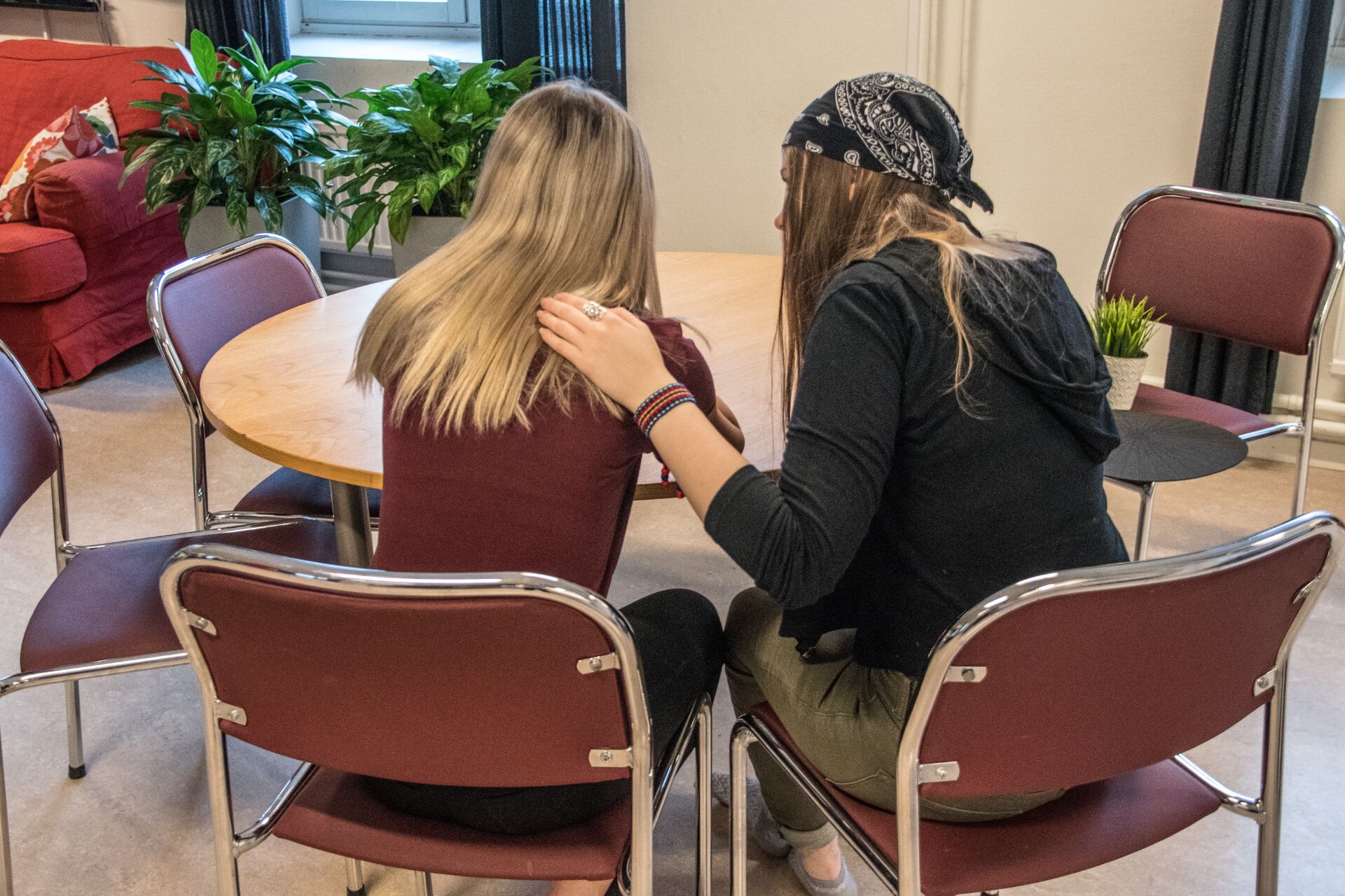 Two girls sitting at a table, one comforting the other, seen from behind.
