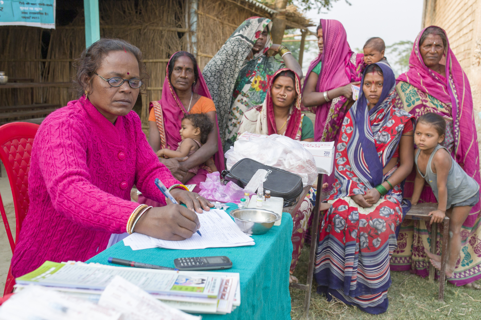 a health worker sits at a desk preparing immunization session a groups of female adults and children sitting and standing waiting