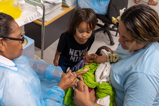 A health worker vaccinates a baby being held by the mother and while a child looks on