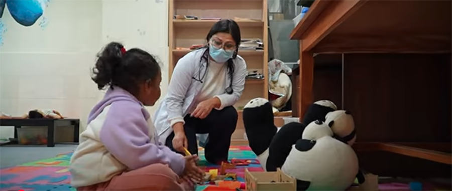 A doctor is kneeling down talking to a young girl sitting in a play area.