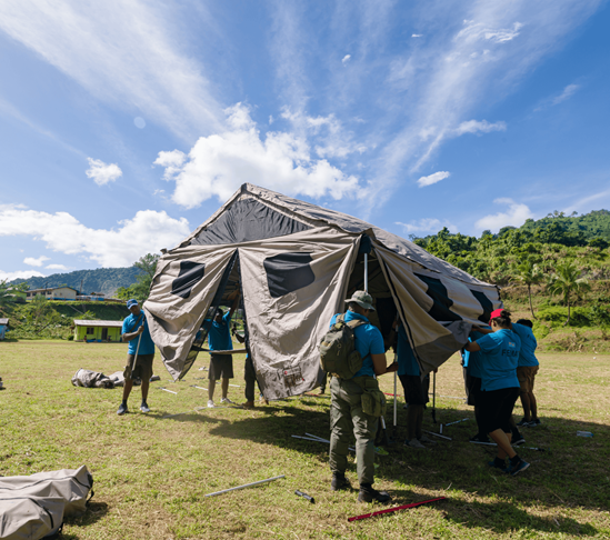 A group of people setting up a tent in a green field under a blue sky