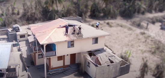 A screenshot of a damaged house after a volcanic eruption. Four people are on the roof.