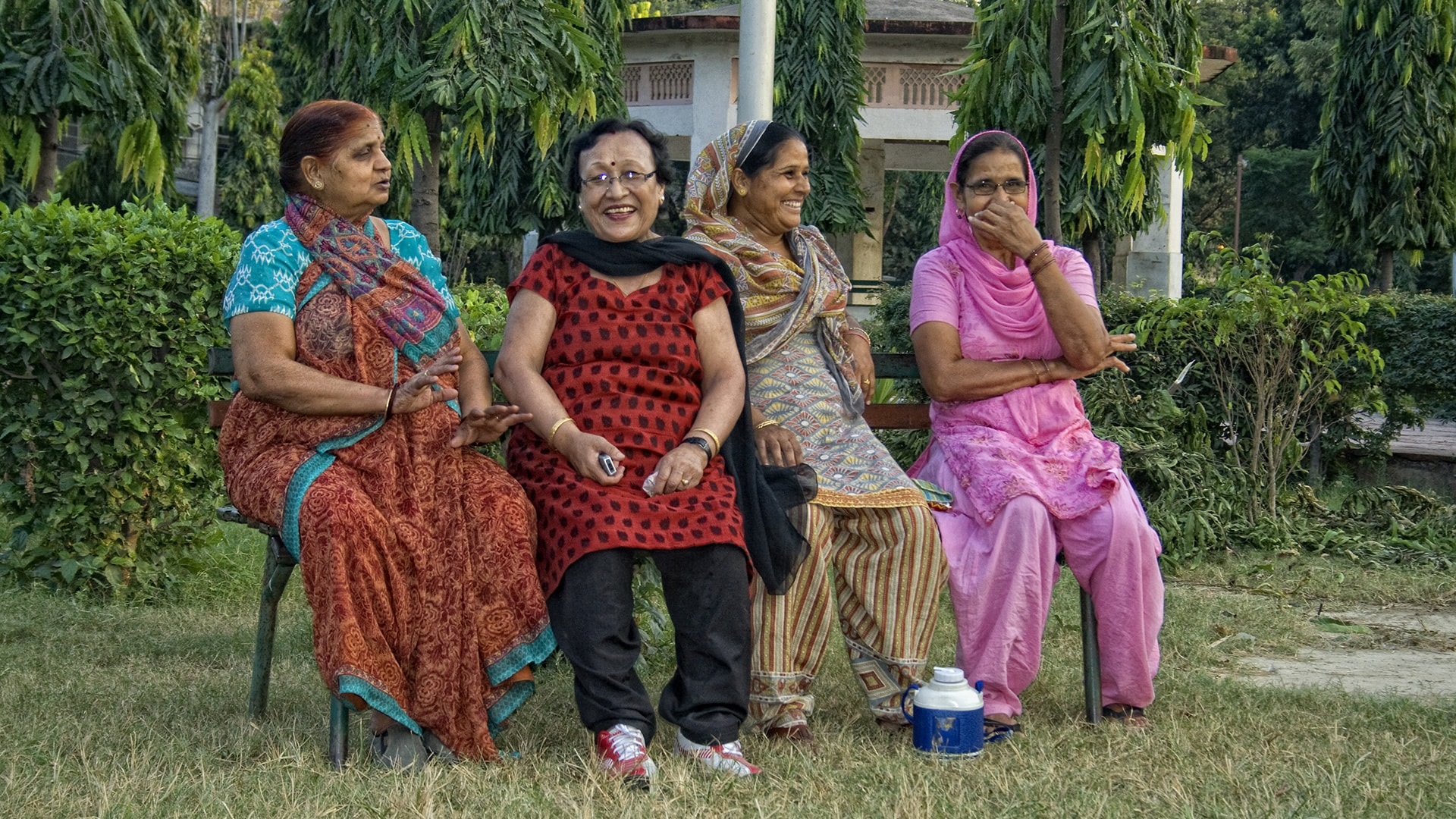 Four older women talk and smile together on a bench in Delhi, India.