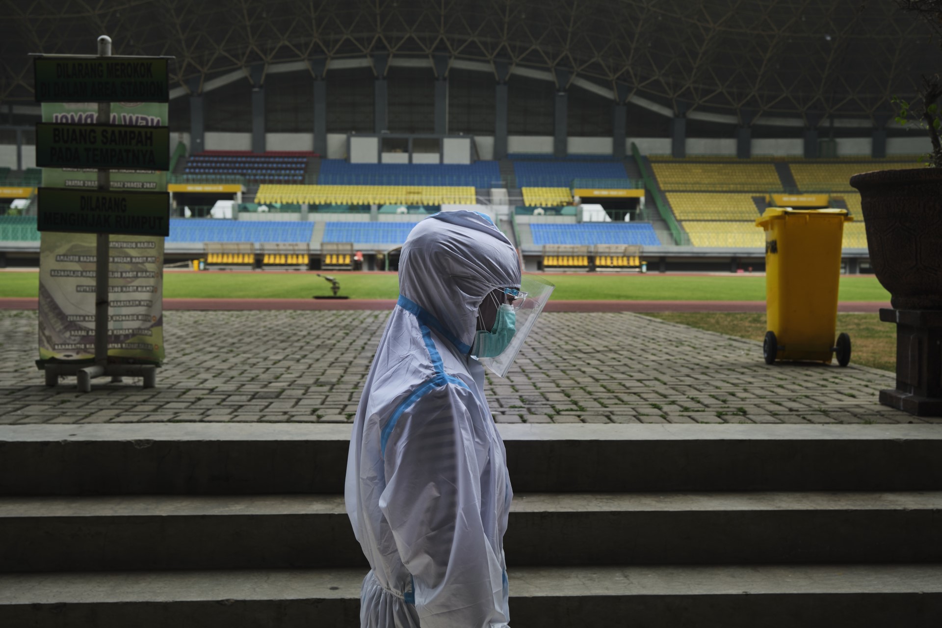 Seorang petugas kesehatan mengenakan alat pelindung diri lengkap dan masker berdiri di stadion kosong dengan tribun berwarna-warni di latar belakang.