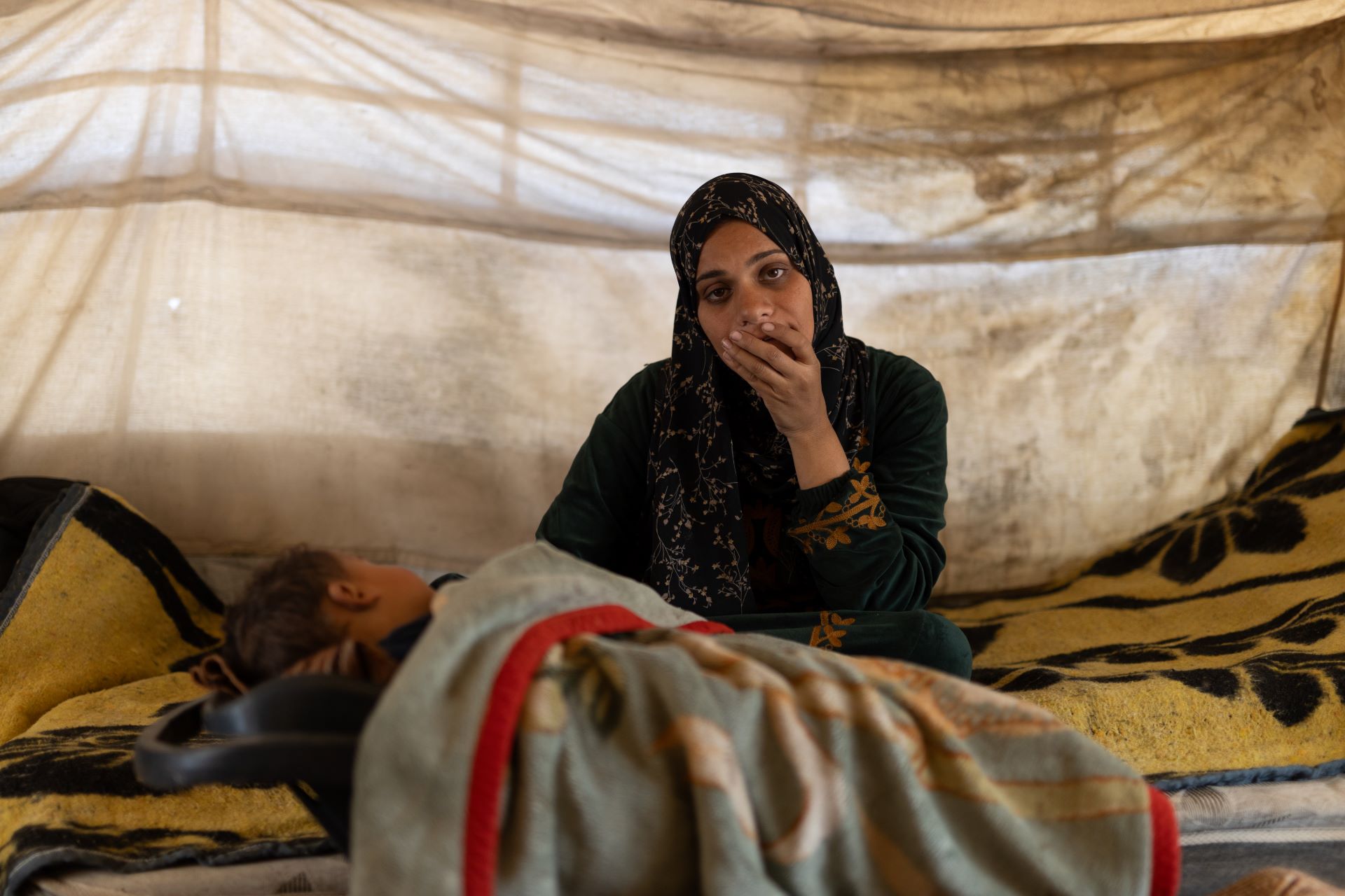 A woman looking distressed with a child laying under a blanket beside her.