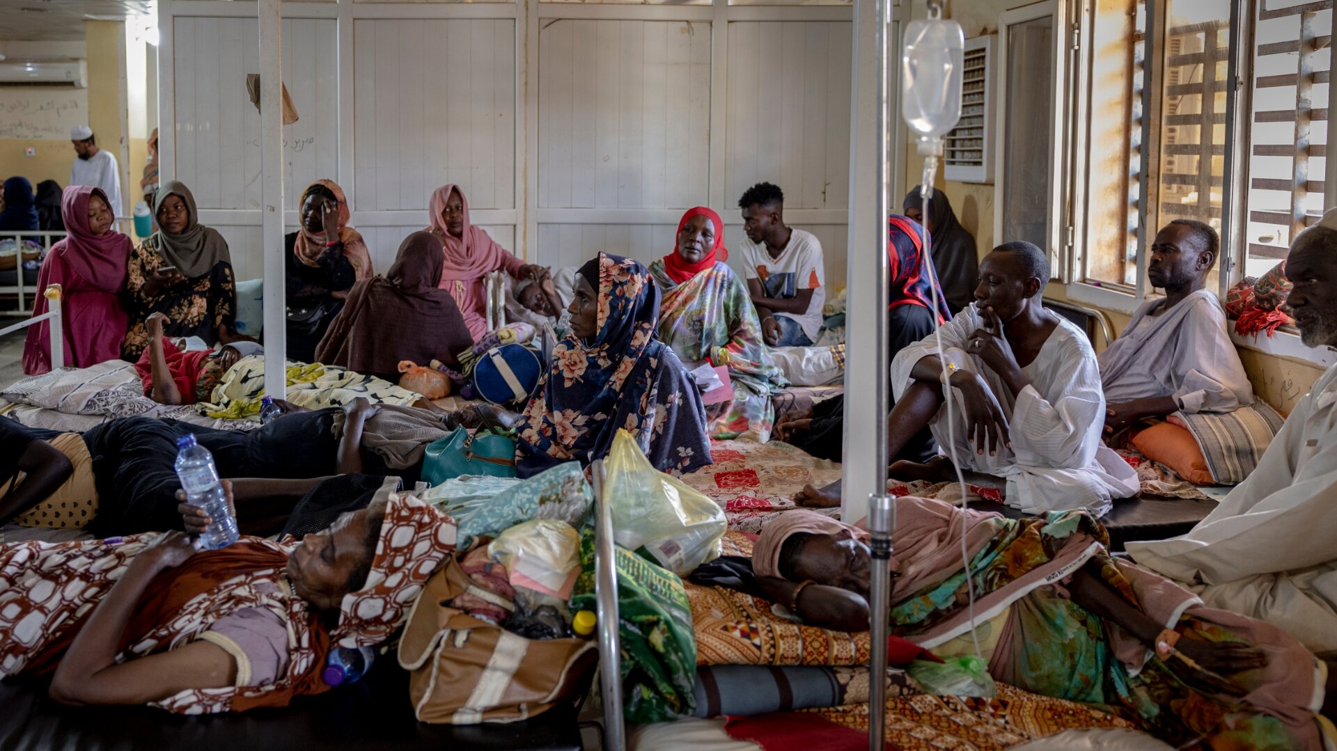 A hospital ward crowded with people, men and women, laying or sitting on beds.