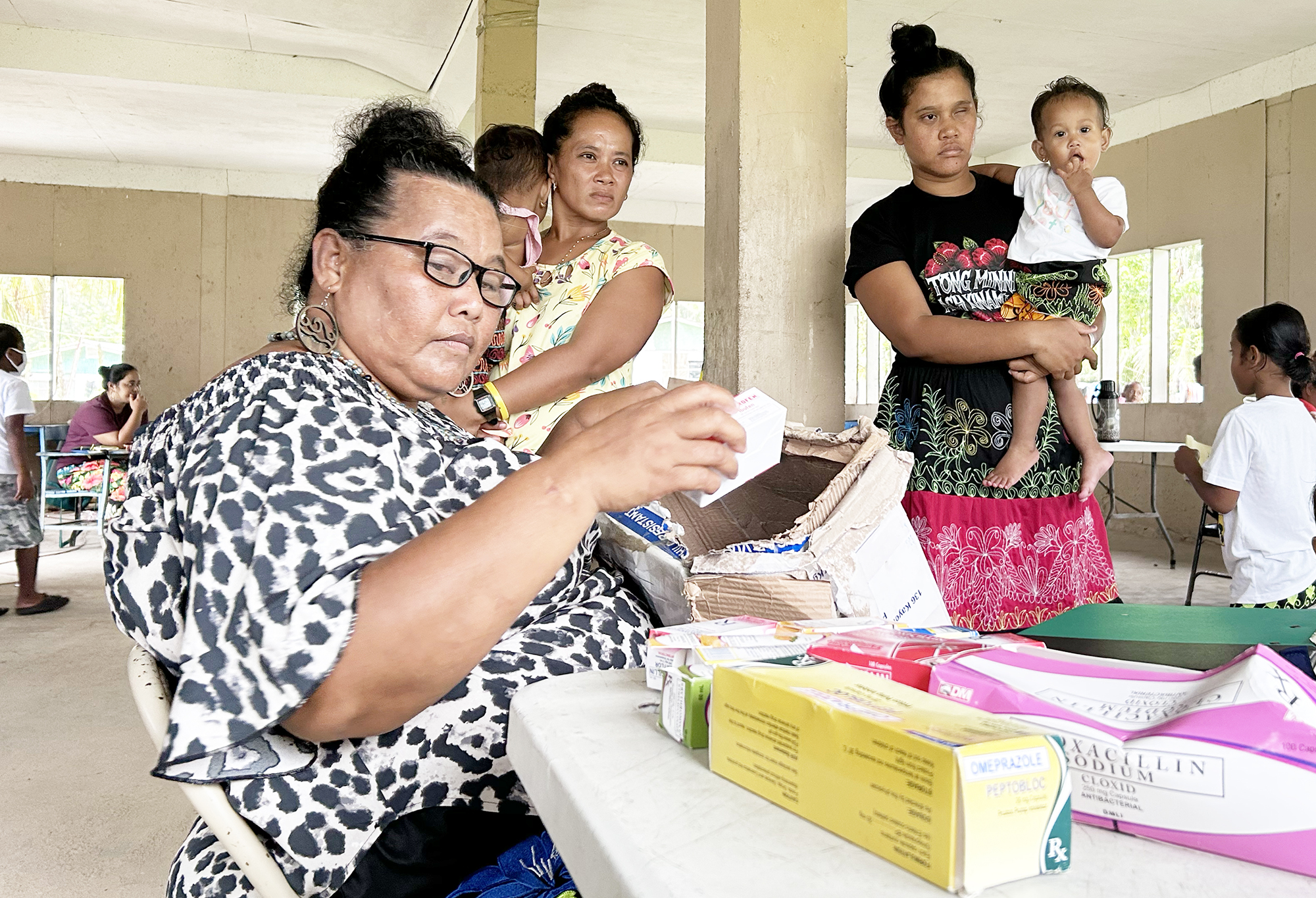 A health assistant examines medicines in the foreground, with two mothers holding their children in the background