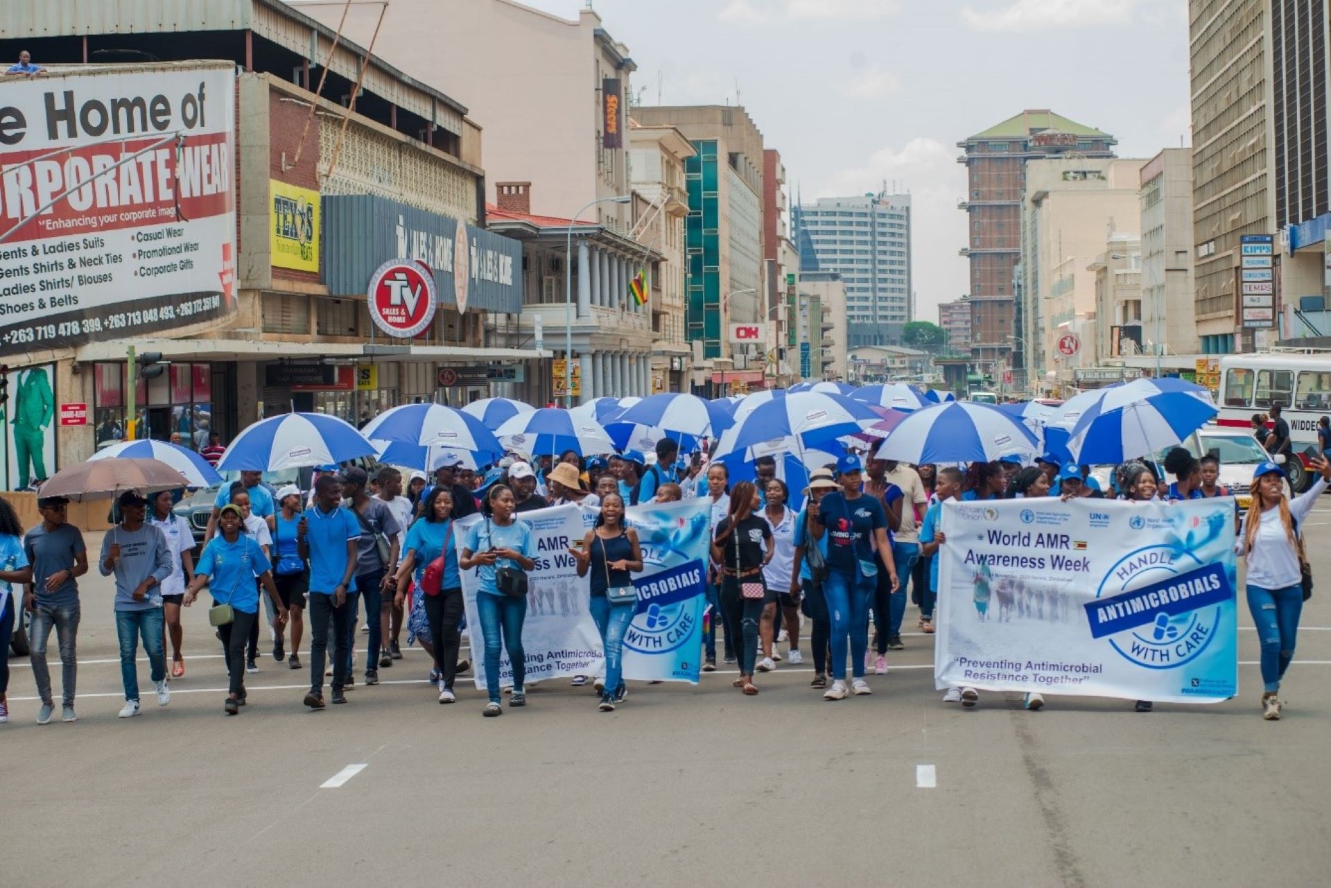 Group of people walking together on the street dressed in blue t-shirts, holding blue umbrellas and the poster with words: "World AMR Awaness Week: Handle antimicrobials with care"