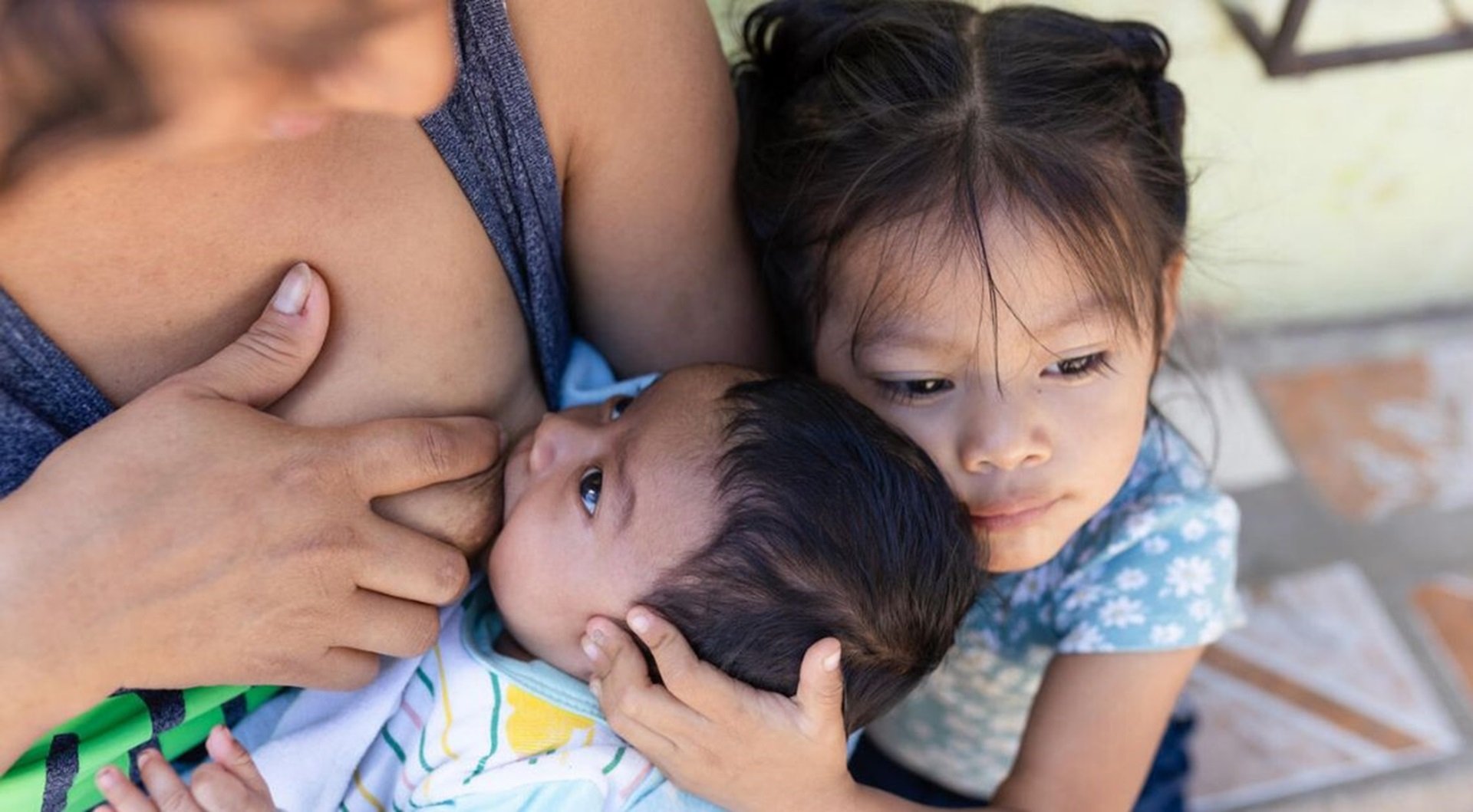 Mother breastfeeding the infant while the older child hugs them.