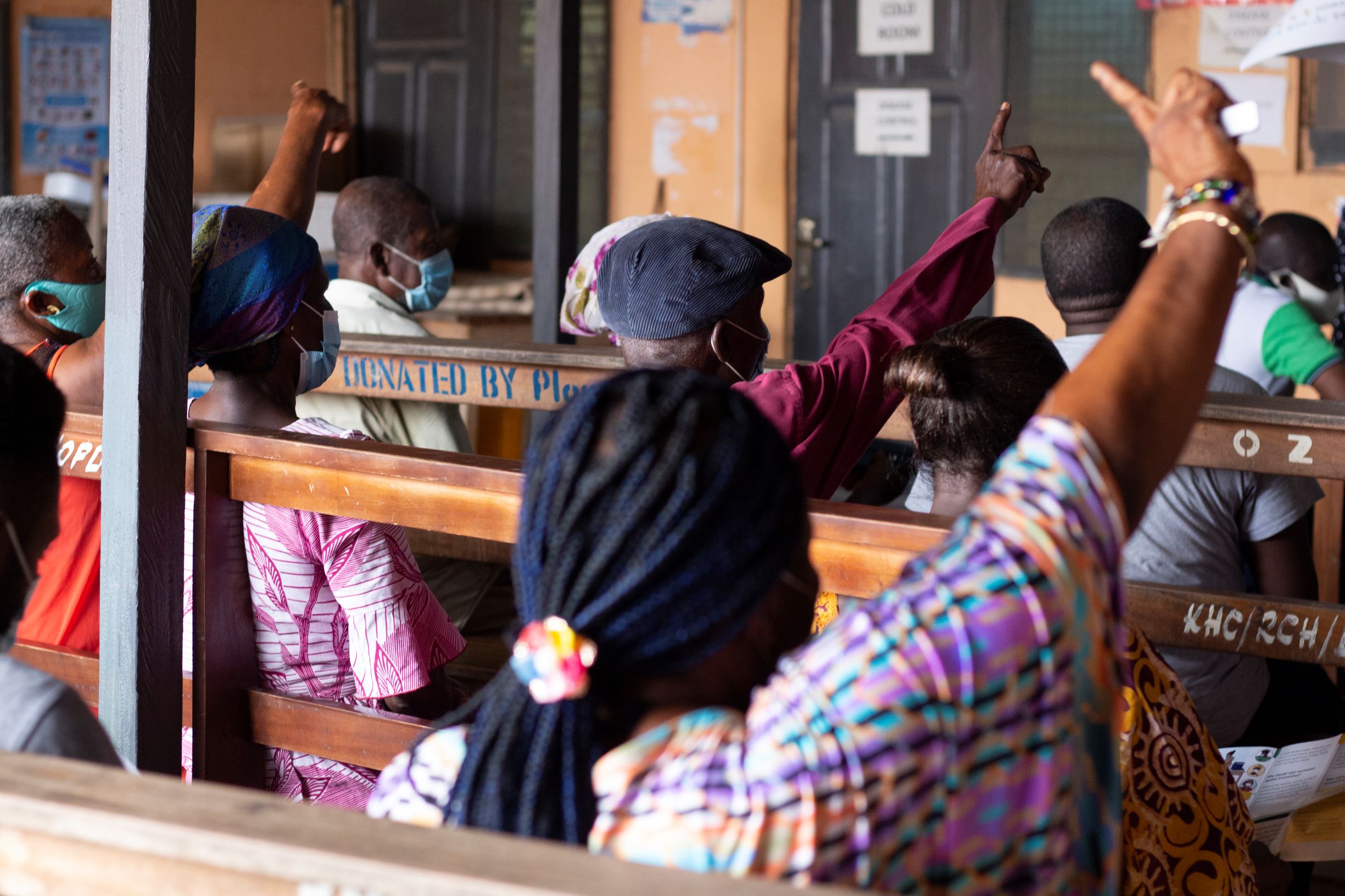 People sitting on wooden benches seen from behind, raising their hands to ask questions.