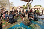 A community gathering led by WHO's Western Pacific Region in a rural setting. Men, women, and children sit in a circle on colorful mats, engaging in a discussion, likely focused on health education or community engagement.