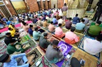 A large group of men in colorful traditional attire, seated on prayer mats inside a mosque, listening to or participating in a prayer service.
