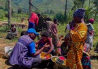 WHO staff member speaks with community members in a rural area, as women wash dishes and children gather nearby.