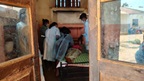 Through a worn wooden doorway, masked healthcare workers in disposable gowns lean over a patient lying on a simple bed.