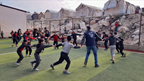 A group of children in a displacement camp hold hands in a circle, playing a game with a facilitator wearing a UOSSM vest on an outdoor sports field. Tents and makeshift shelters are visible in the background.