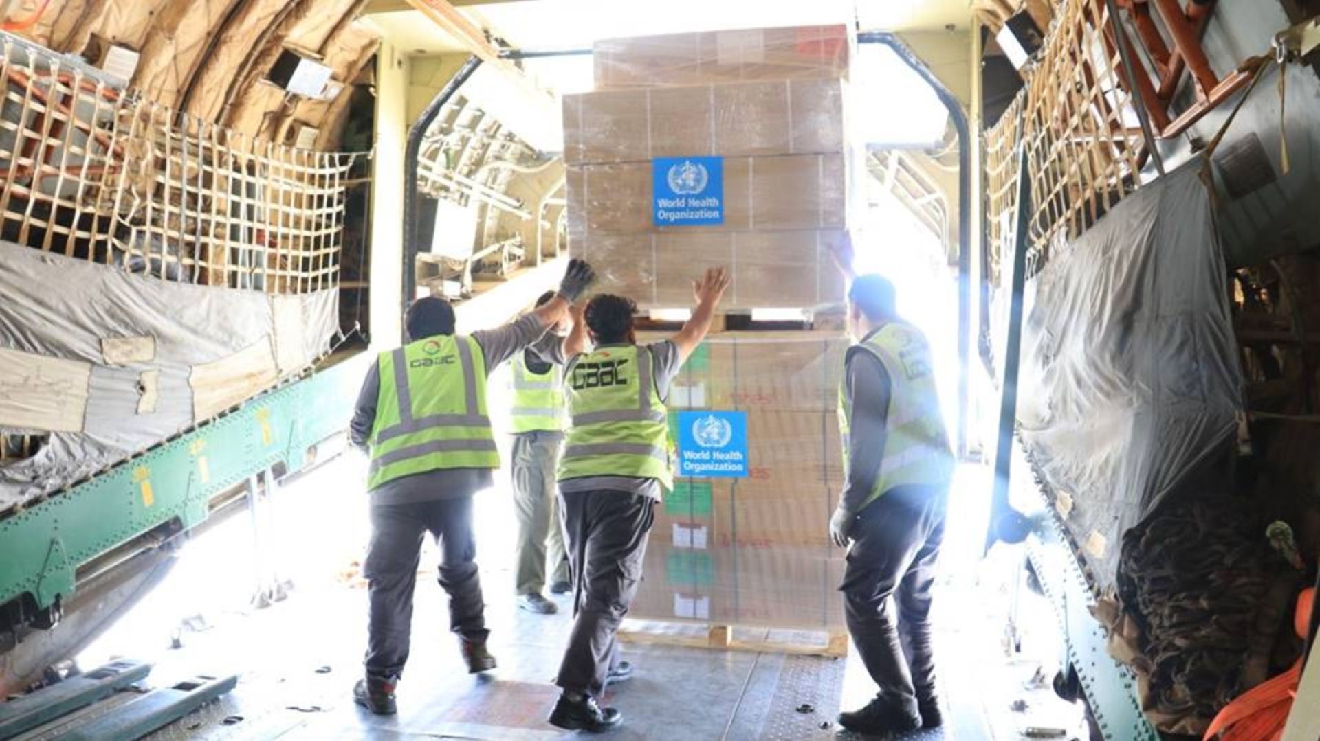 The image shows workers unloading large boxes marked with the World Health Organization logo from an aircraft.