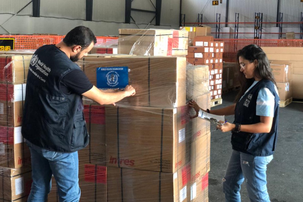 Two people in WHO vests checks boxes in a warehouse.
