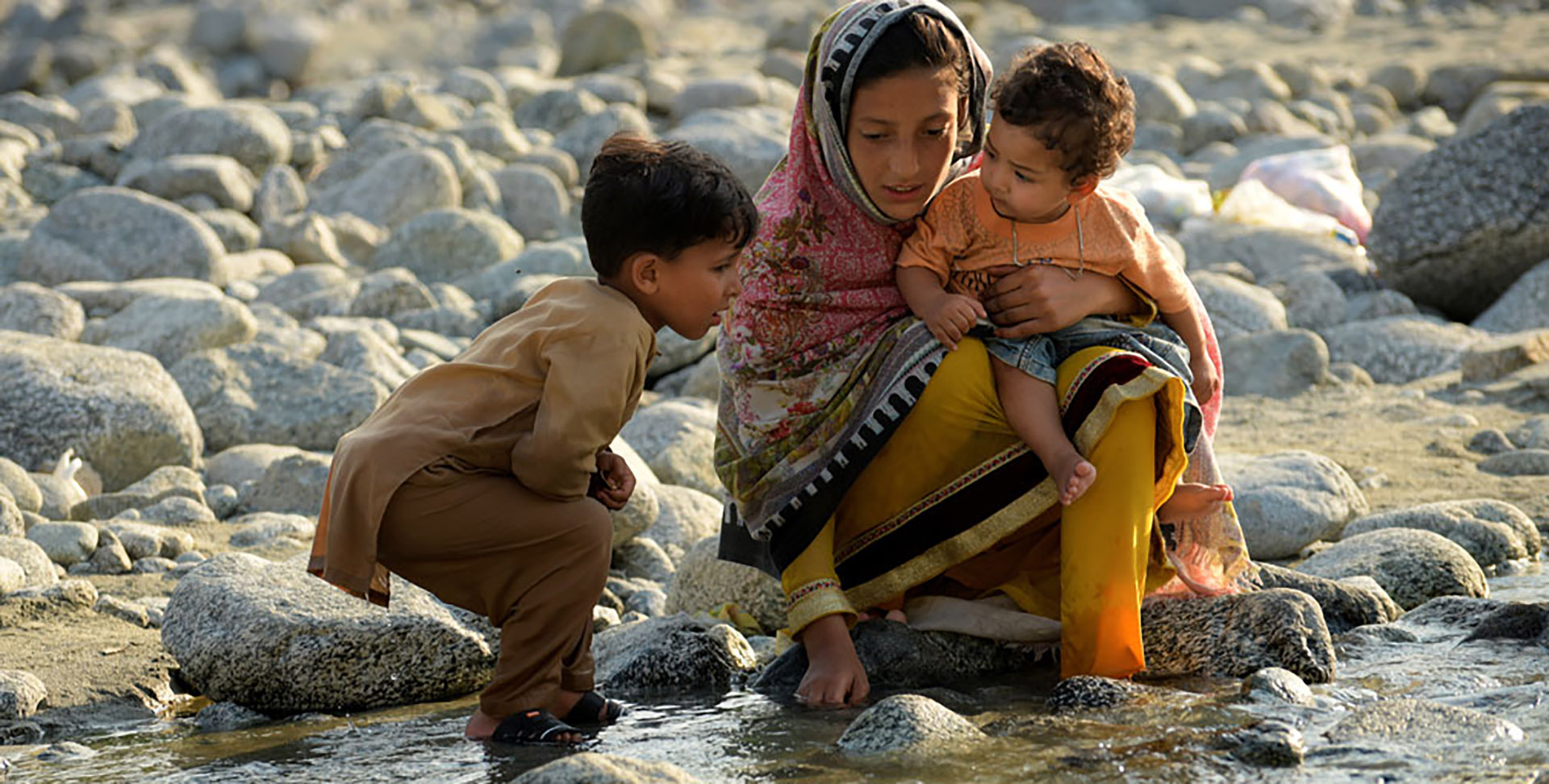 A mother sitting on rocks holding a small child on her lap with a young child looking on