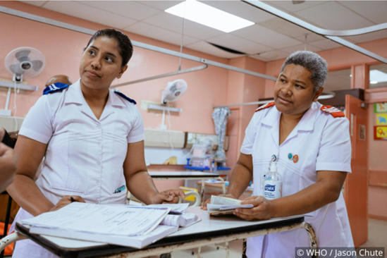 Two healthcare professionals in white uniforms discuss patient files at a desk in a medical facility.