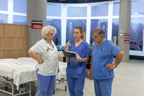 Three health professionals review a clipboard in a hospital training room with beds in the background.