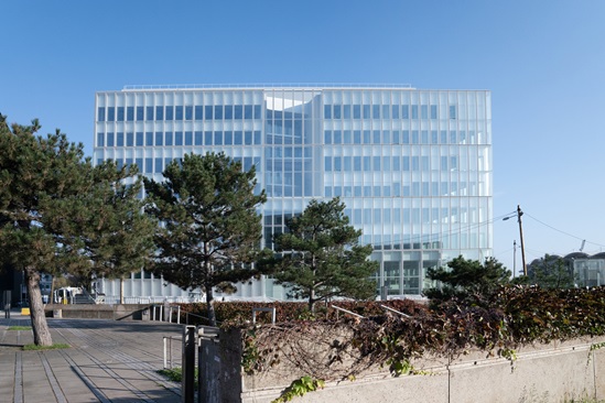 Un grand bâtiment moderne avec une façade en verre, entouré de quelques arbres et d'une haie.