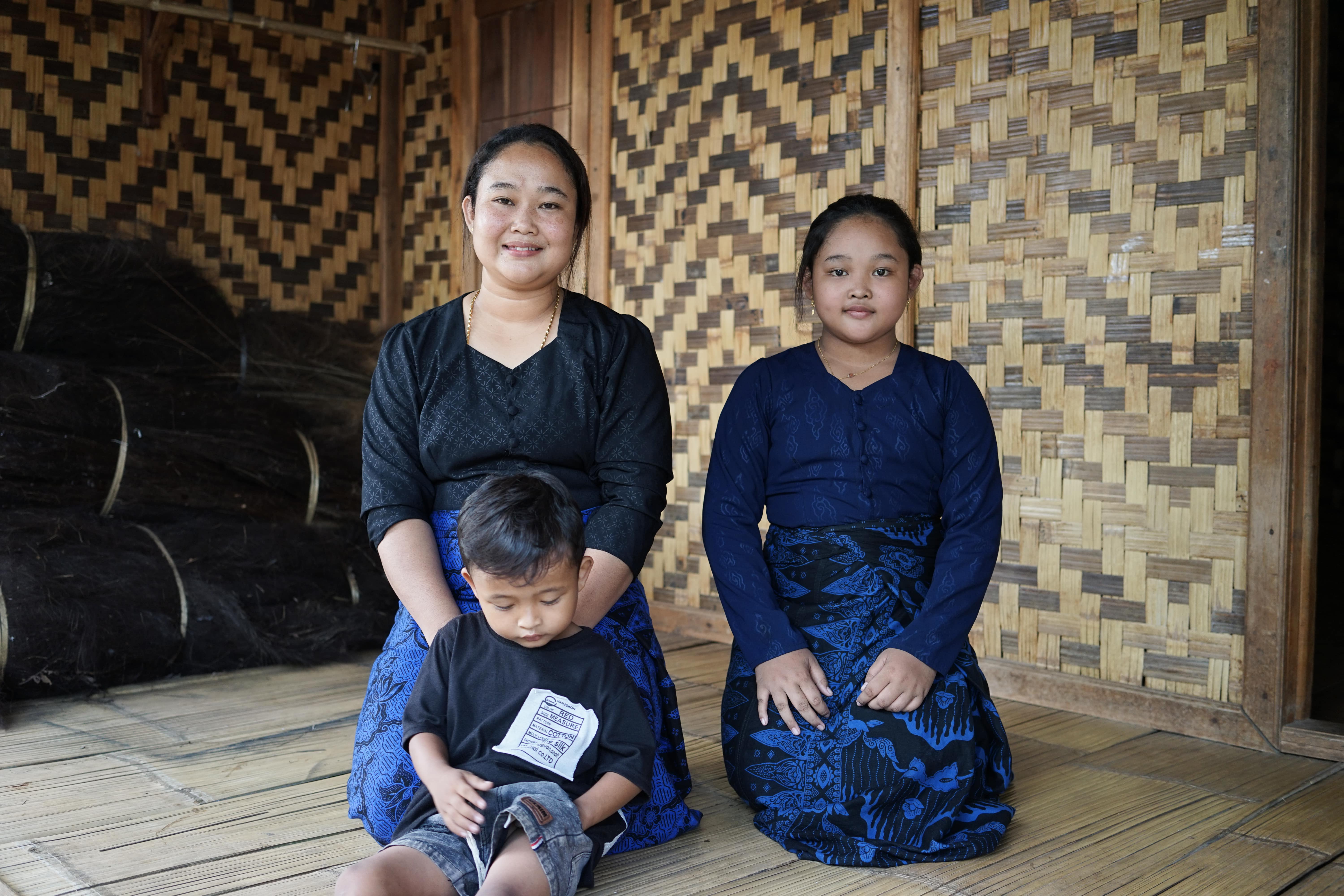 A mother, her daughter, and her small son sitting on a wooden floor against a backdrop of woven bamboo walls. The adult woman wears a black and blue kebaya, while the young girl wears a dark blue kebaya.