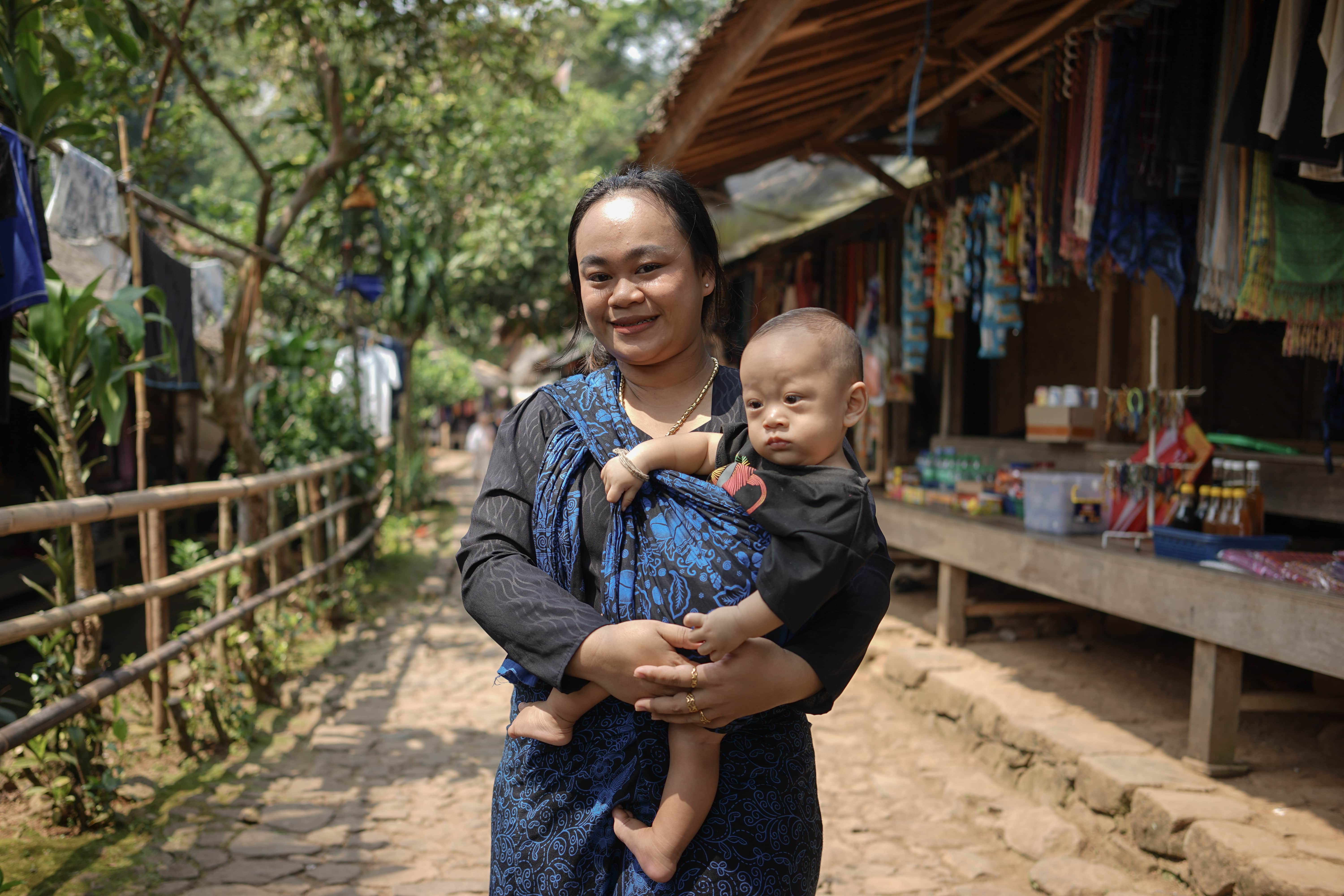 A woman wearing a black and blue kebaya holding a baby in front of a traditional shop. The background shows a village path with wooden houses, trees, and a peaceful rural atmosphere