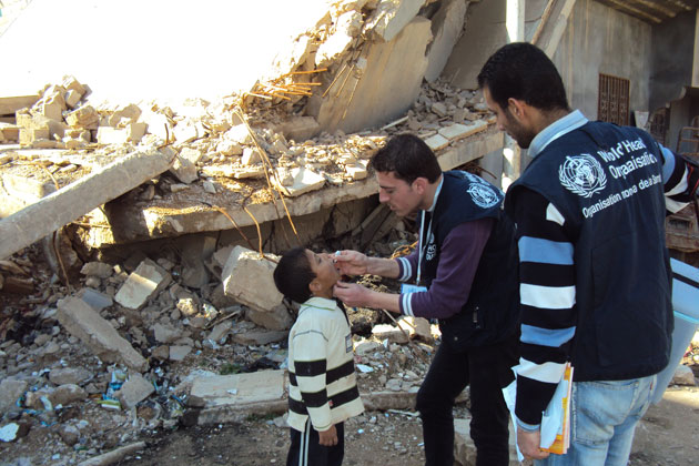 Two men with WHO vests give an oral vaccination to a child, in front of a collapsed building.