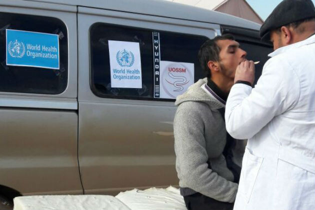 A doctor examines the throat of a man sitting on a stretcher next to an ambulance.
