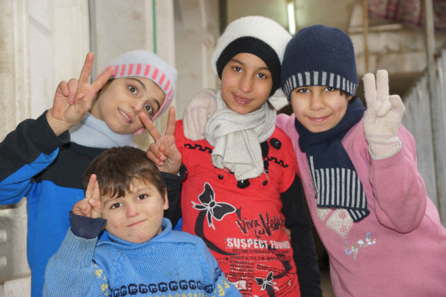 Four smiling children wearing toques and scarves each hold up 2 fingers in a peace sign.
