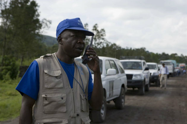 A man standing in front of a row of vehicles speaks into a radio.