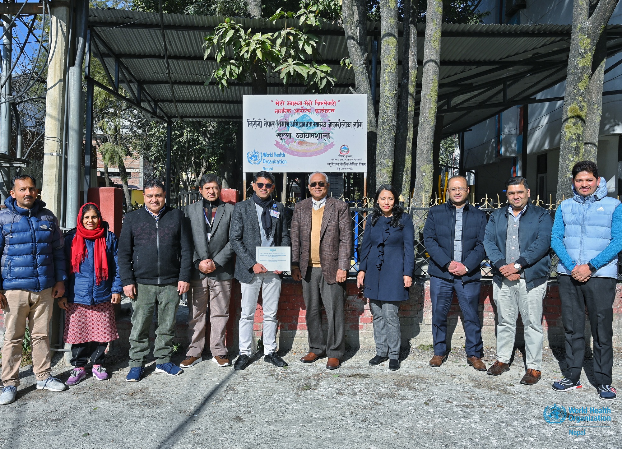 A group image where WHO Representative to Nepal is handing over the certificate of gym equipment to Department of Ayurveda and Alternative Medicines in front of one of the sites where the equipment were installed