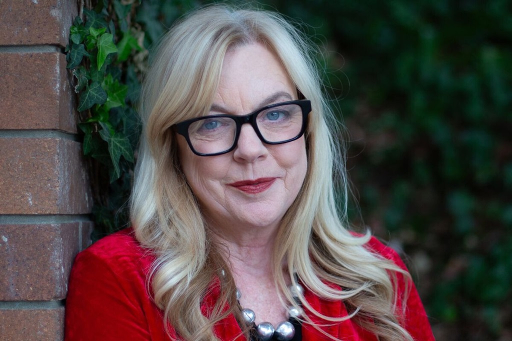 Portrait photo of a woman, wearing a red jacket and glasses, leaning against a brick wall.