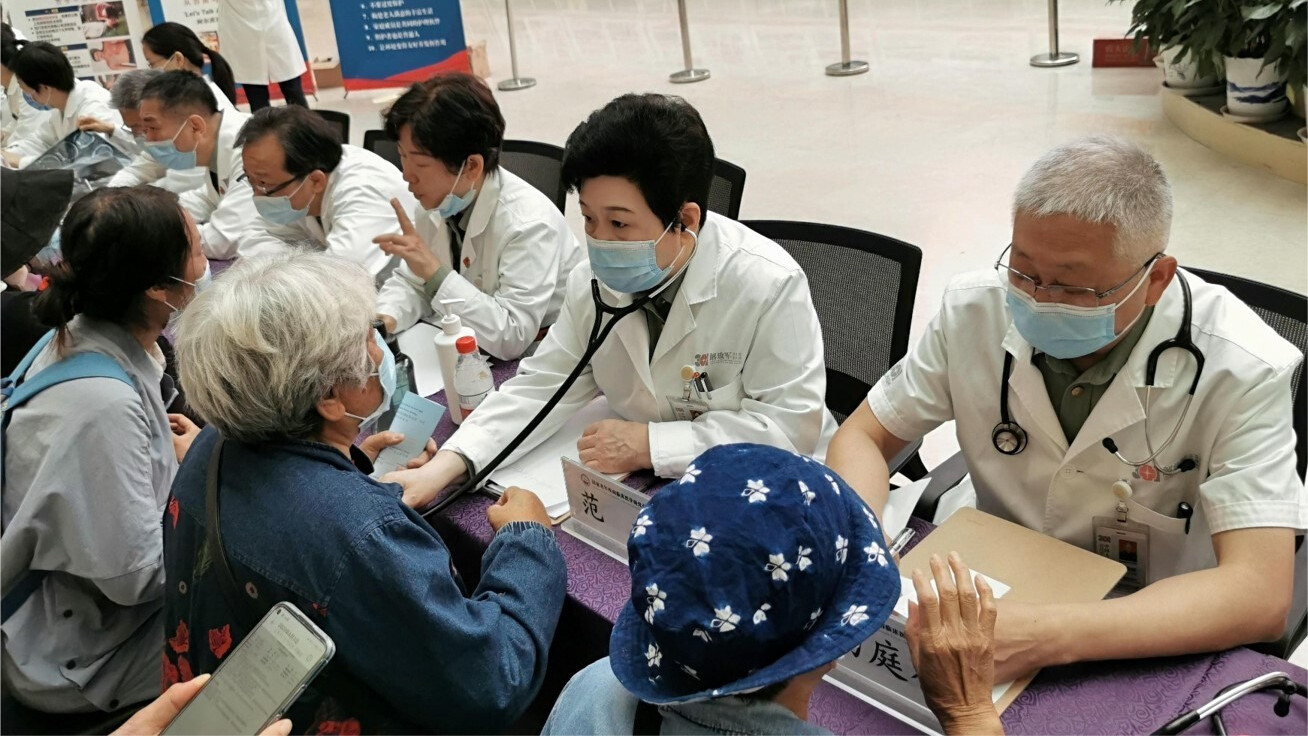 Doctors wearing face masks sits on one side of a long table attending to elderly people sitting in front of them on the other side of the table.