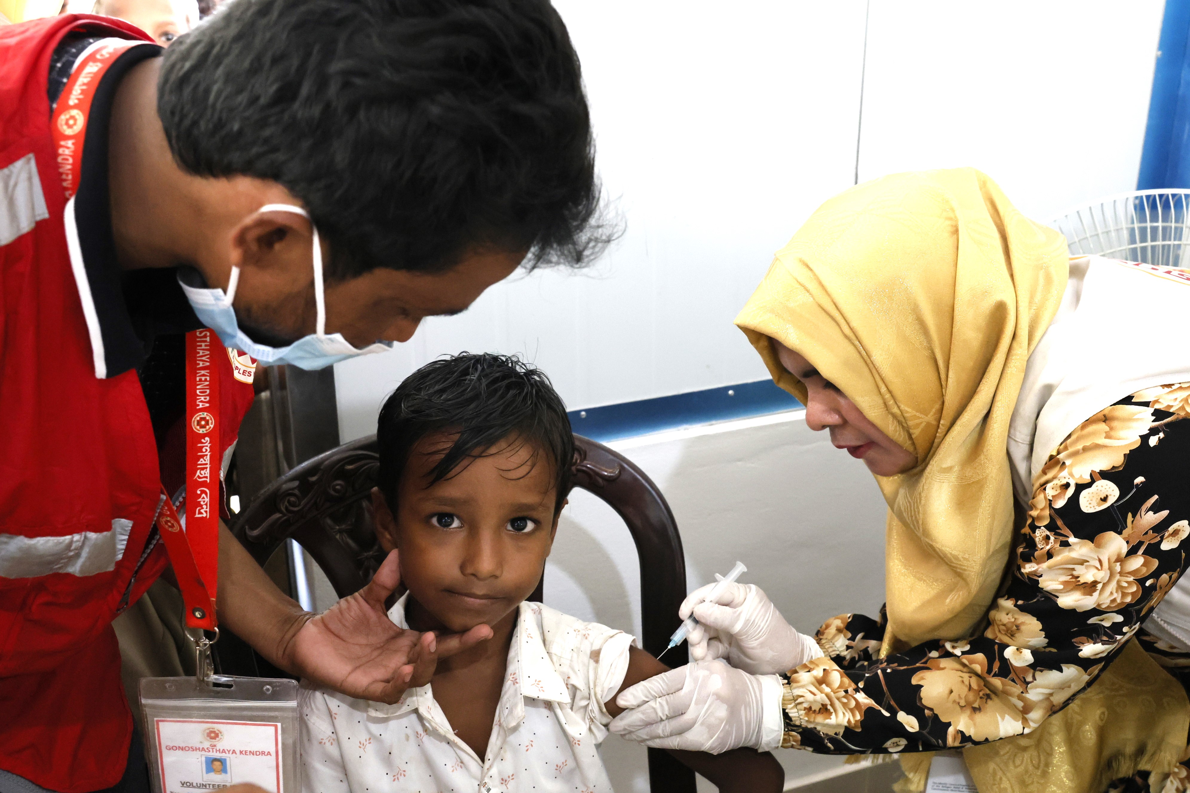 A female vaccinator administers TCV to a Rohingya child, with volunteers supporting