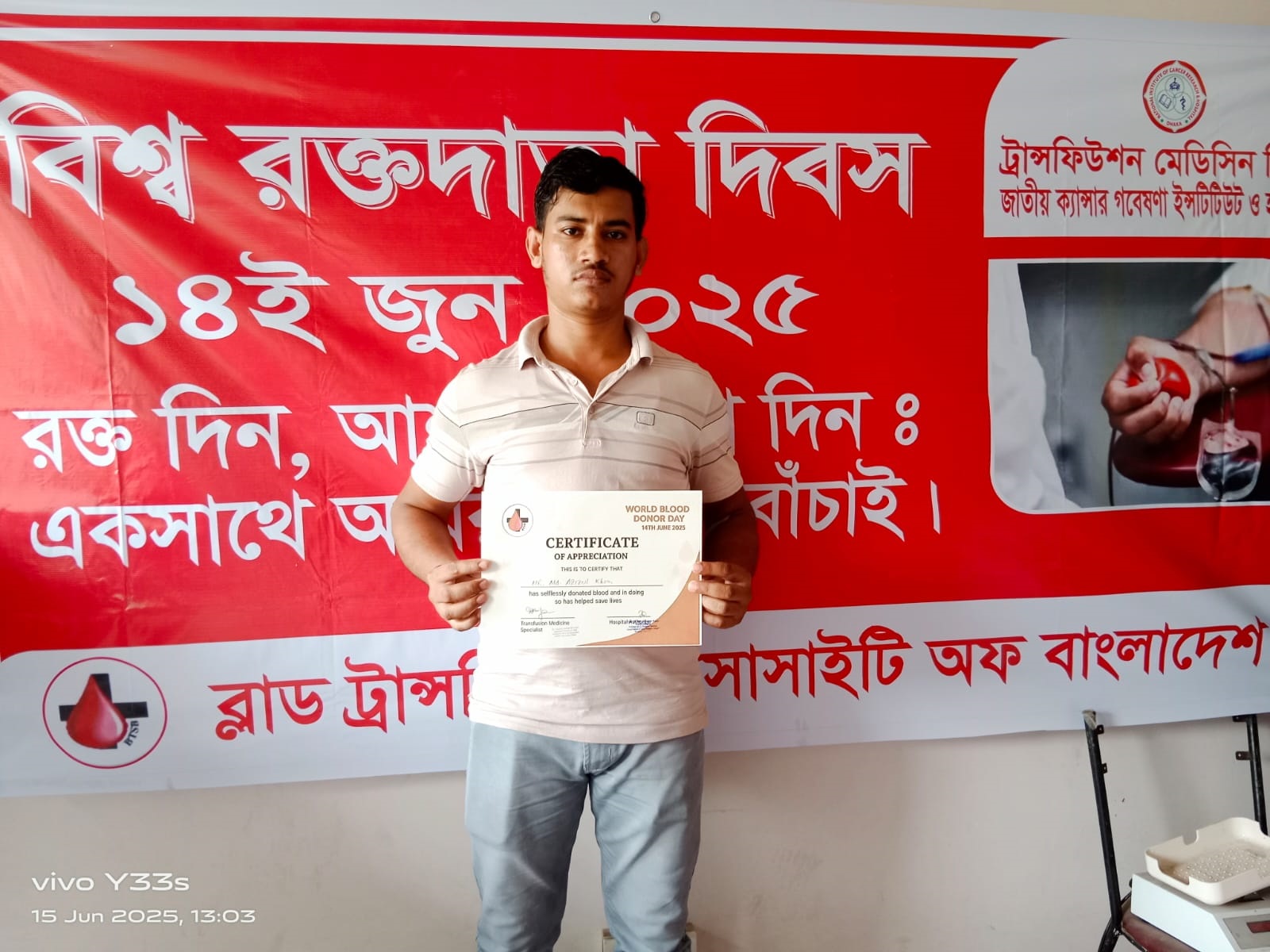 A man standing and holding a certificate in front of a red banner for World Blood Donor Day, with Bangla and English text.