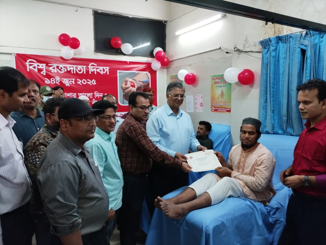 A man sitting on a hospital bed receives a certificate from officials during a World Blood Donor Day event, with red-white balloons in the background.