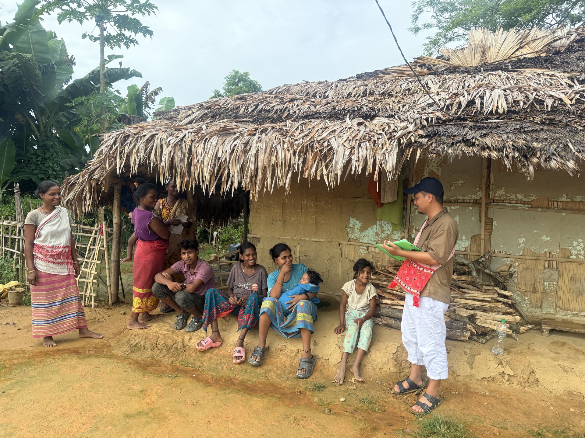 A community gathers outside their traditional thatched-roof hut in a rural area, engaging in daily activities.