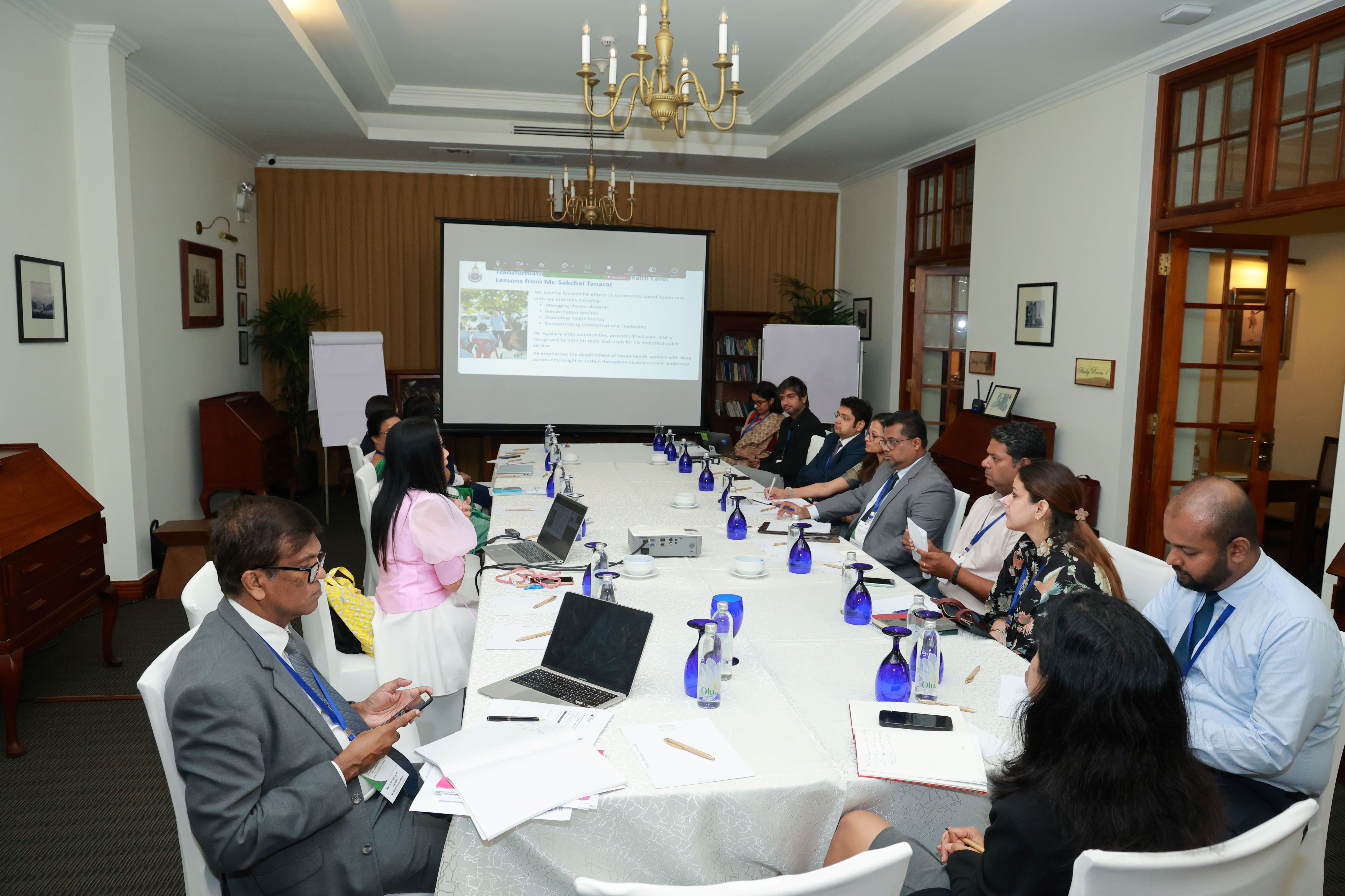 People in a meeting room around a long table, viewing a presentation on a large screen.