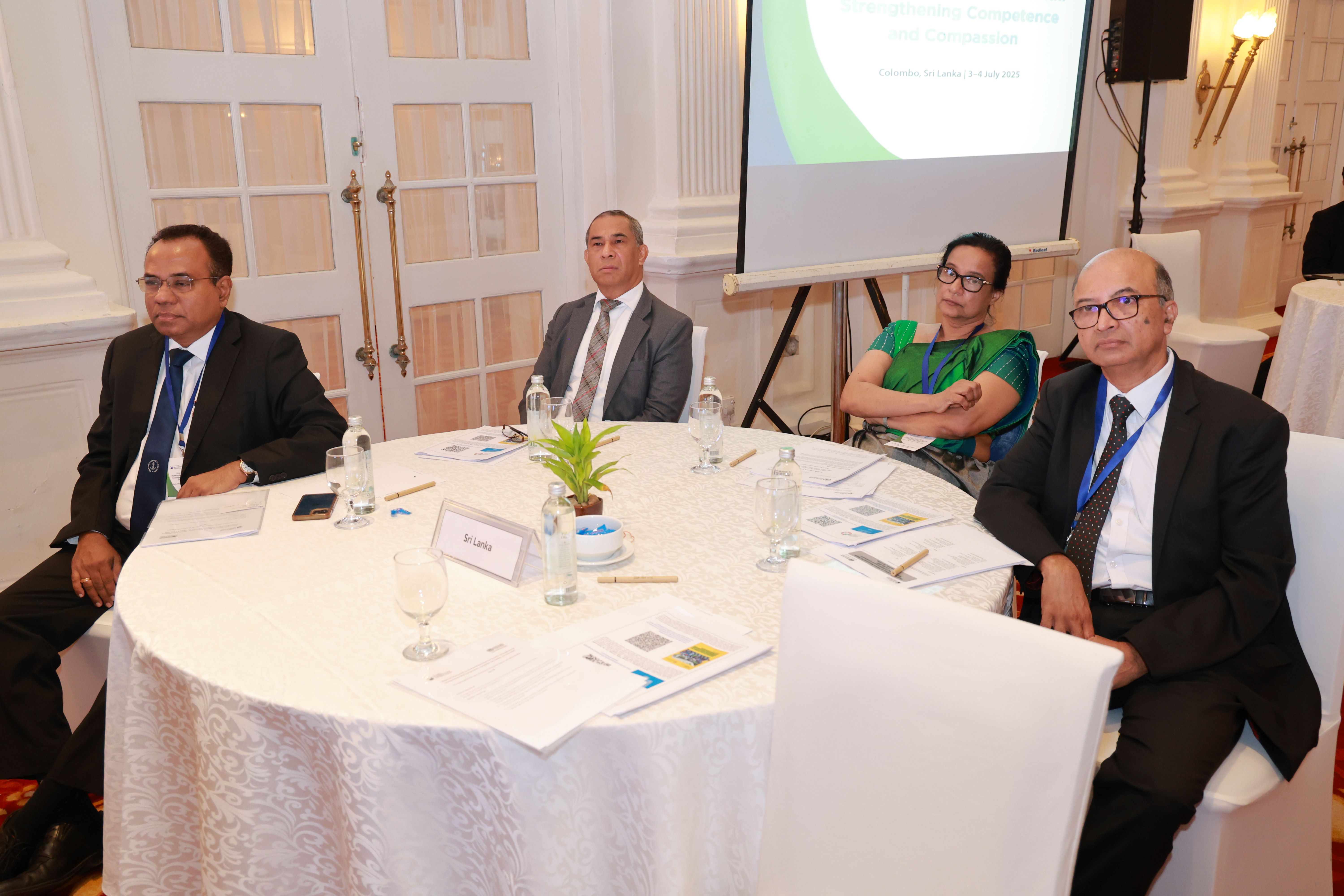 Four people seated around a round table in a formal meeting, with papers, water bottles, and a screen in the background.