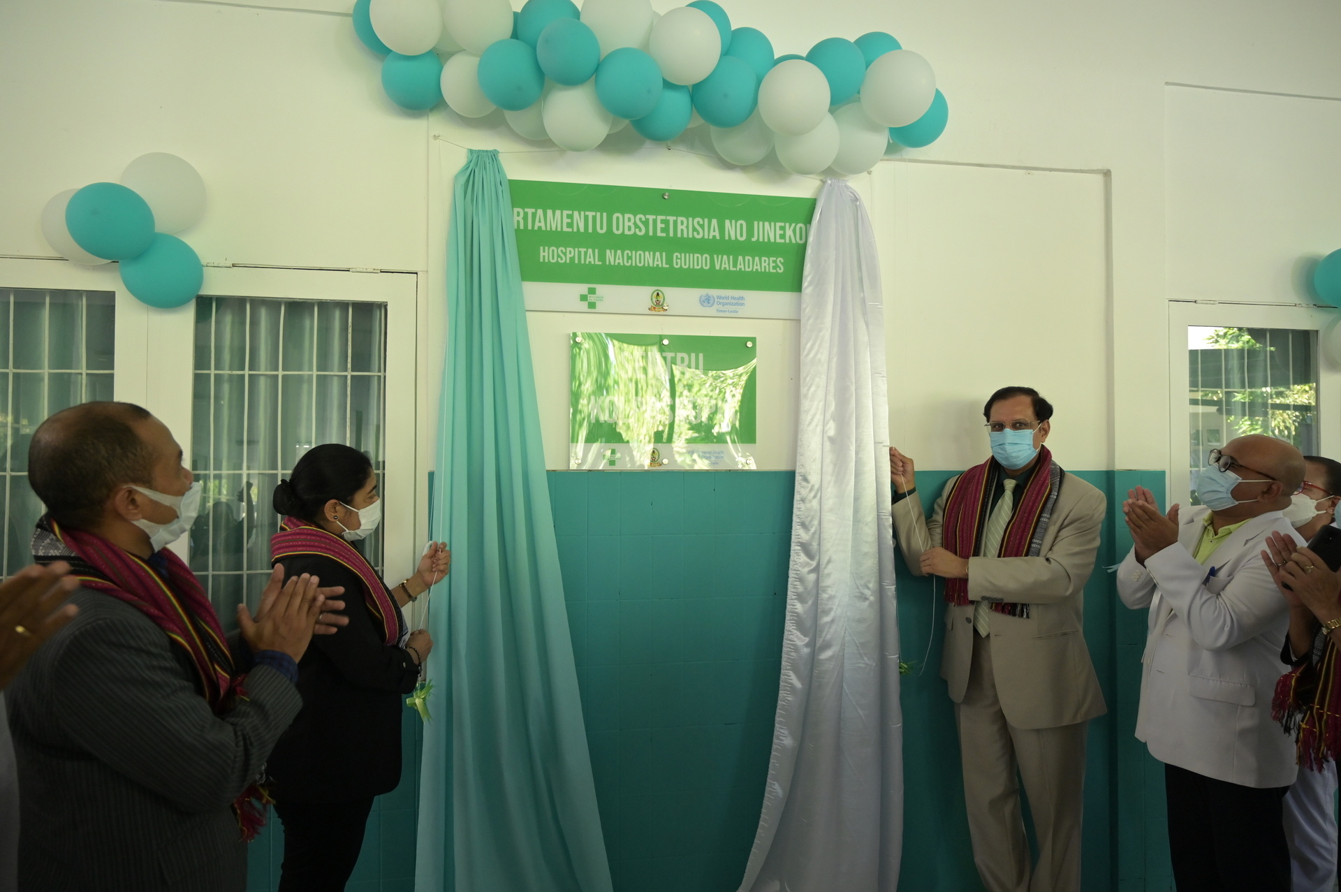 Five people wearing masks, two of them are opening one blue and one white curtain