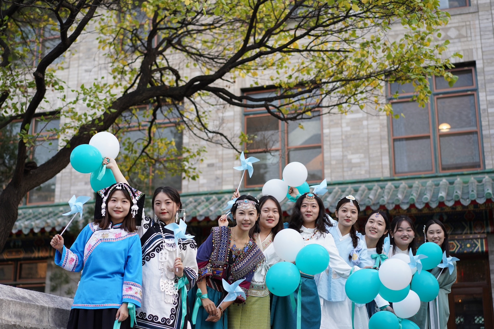 A group of students at Peking Union Medical College in Beijing stand outdoors holding teal and white balloons and pinwheels under a leafy tree, celebrating World Cervical Cancer Elimination Day.