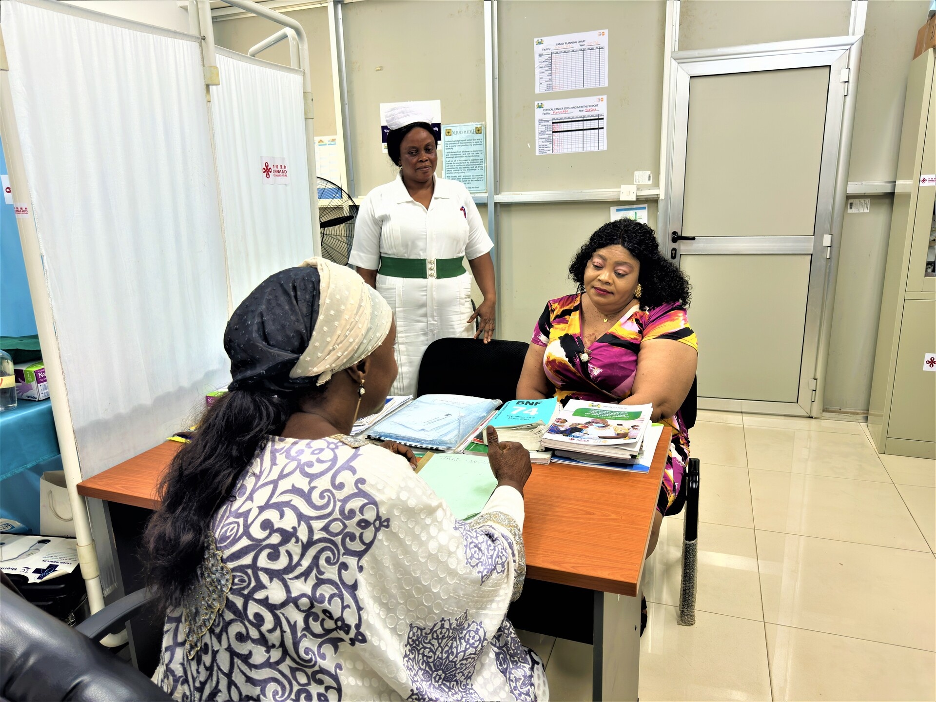 A health worker is speaking to a female patient sitting at a desk with a nurse standing behind.