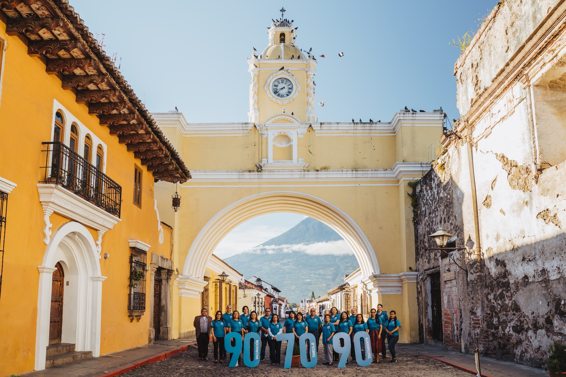 A group of people in teal shirts stand beneath the iconic Santa Catalina Arch in Antigua, Guatemala, holding large “90-70-90” letters to mark the Day of Action for Cervical Cancer Elimination as part of the SUCCESS Project.
