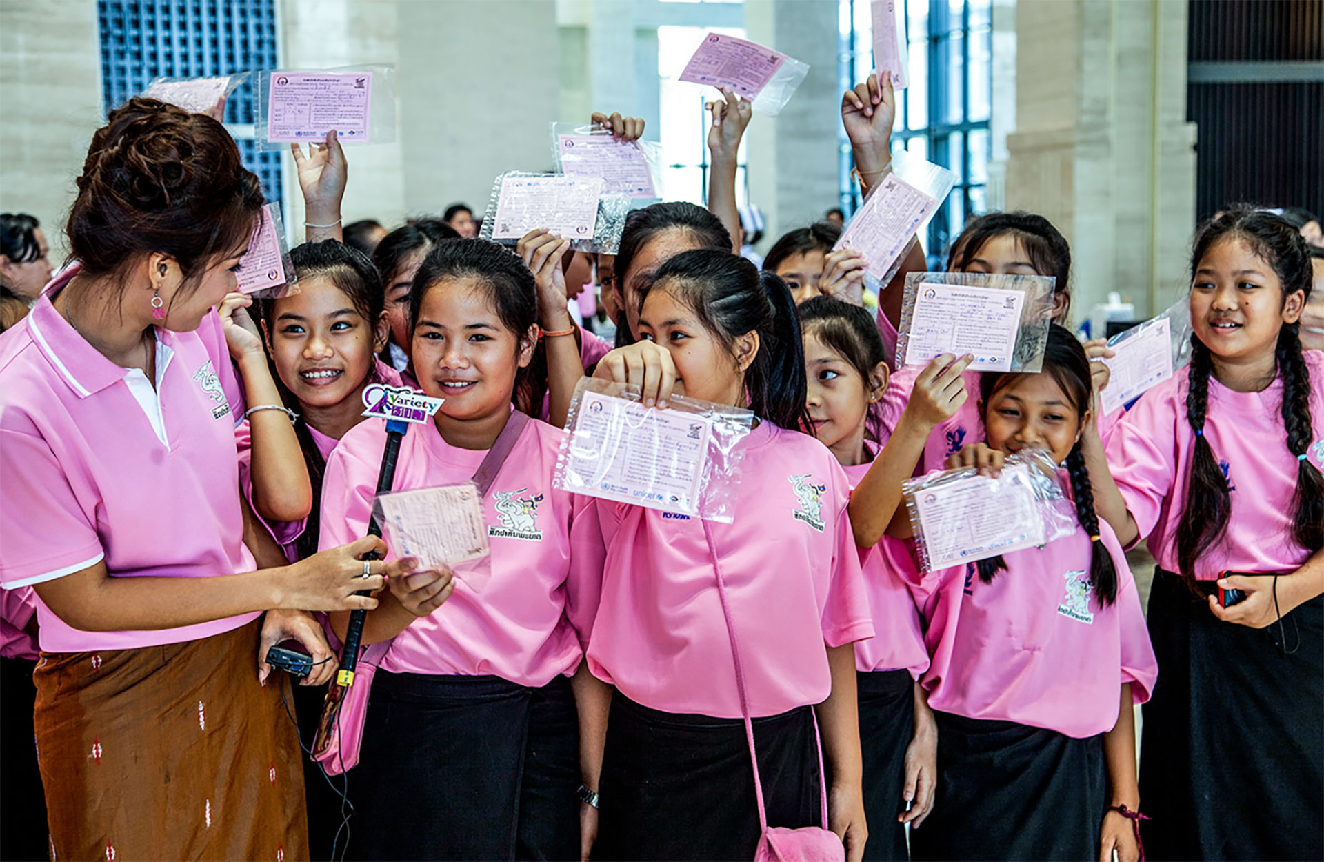 A group of young girls dressed in pink tops holding vaccination cards one girl is being interviewed by a woman