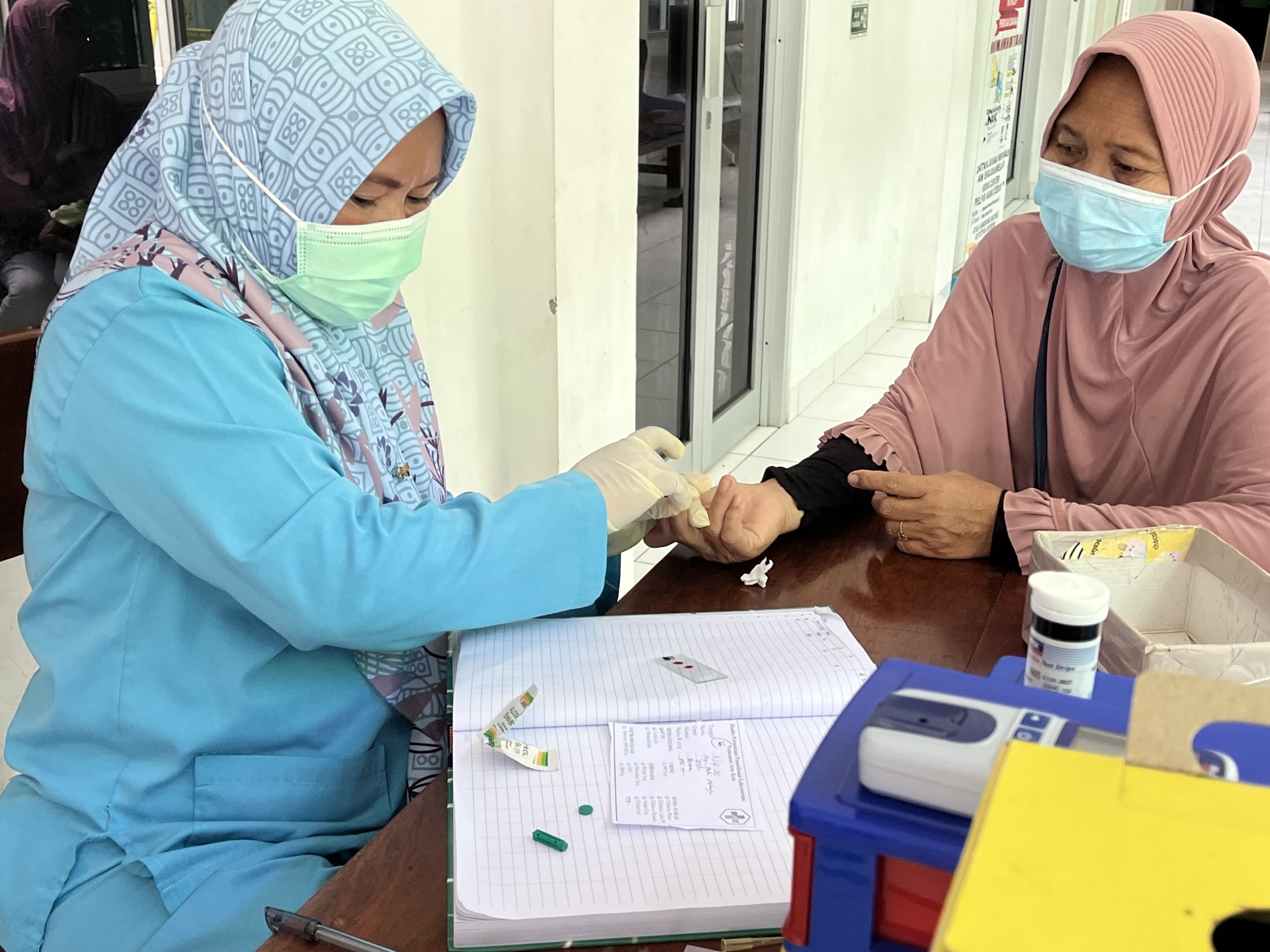 Two women in hijabs sitting across a table in a primary health centre. One is a health care worker who is performing blood test on another woman's finger.