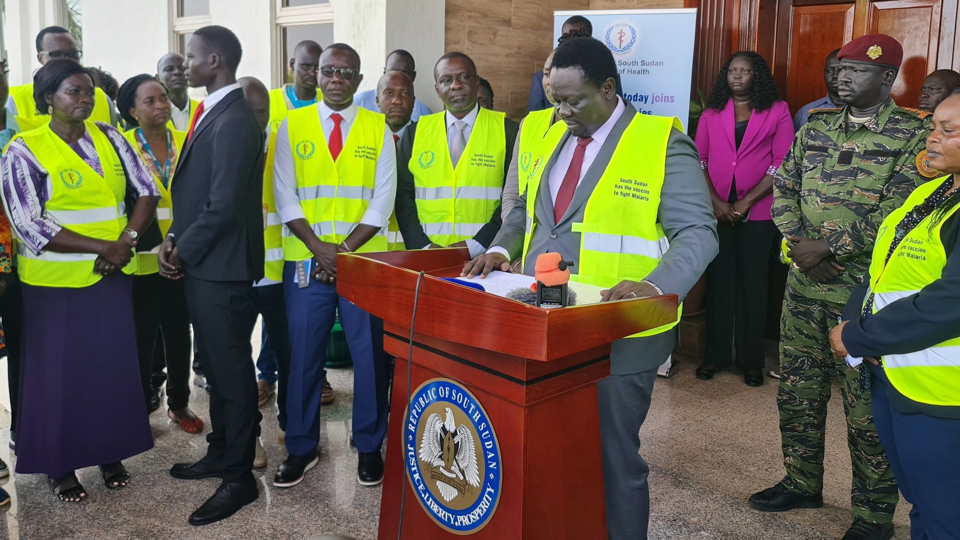 In the centre of the image there is a man standing at a speaker's podium wearing a yellow alert vest, around him there are people standing in a semi-circle, also wearing yellow alert vests.