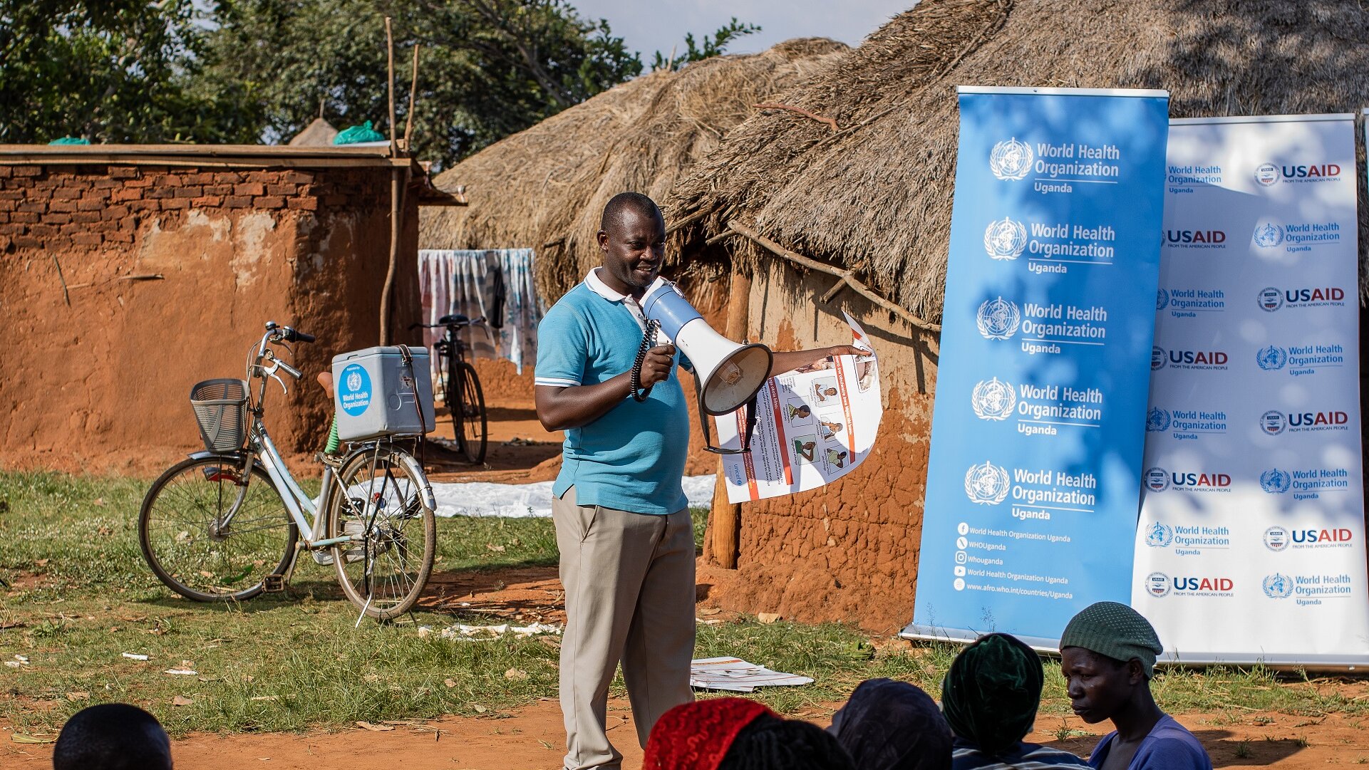 Mpox community engagement Uganda A community health extension worker, standing next to two banderols with WHO and USAID logos, is talking to a group of people using a megaphone.
