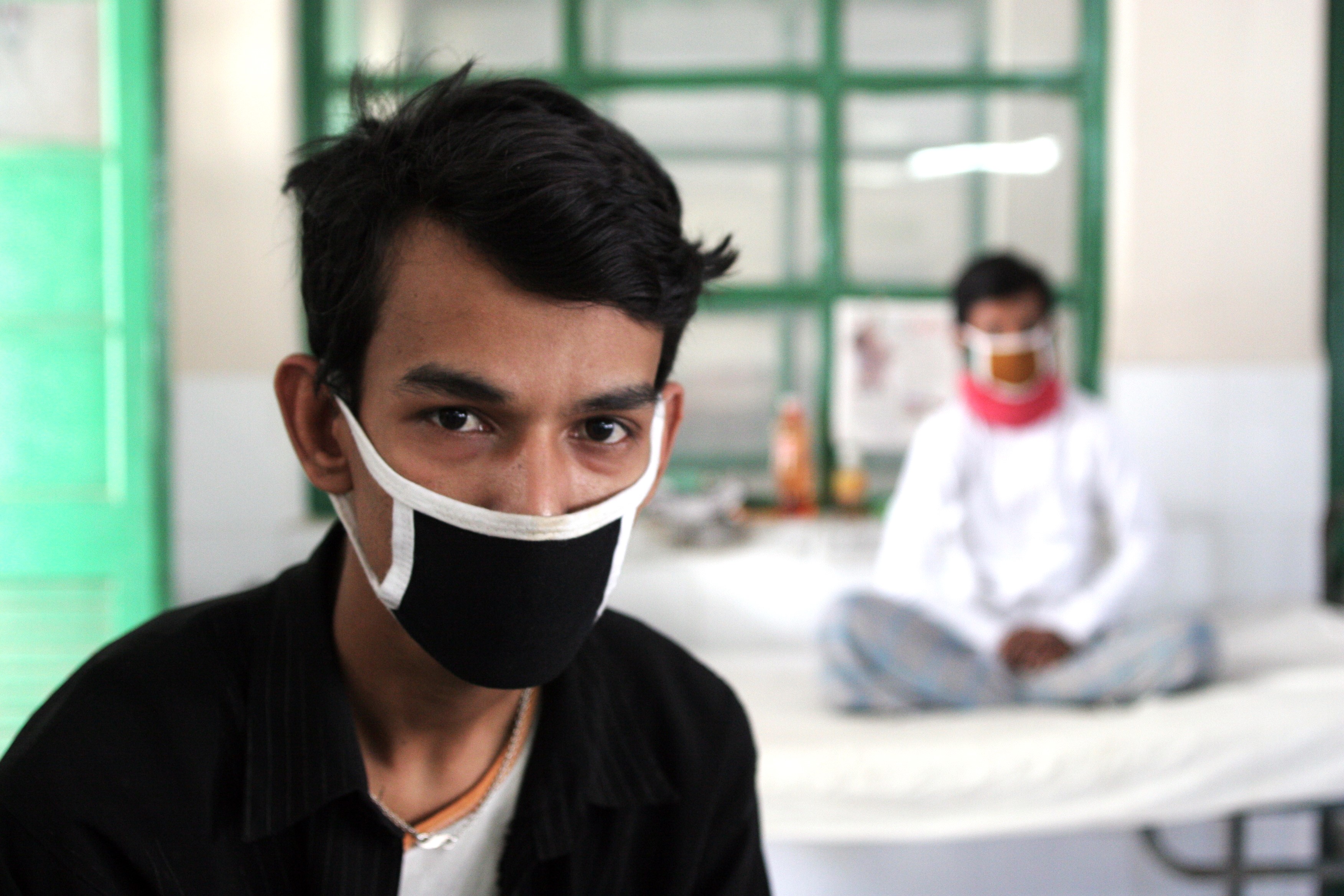 a patient on bed with mask in background and a man in foreground with black mask and black shirt