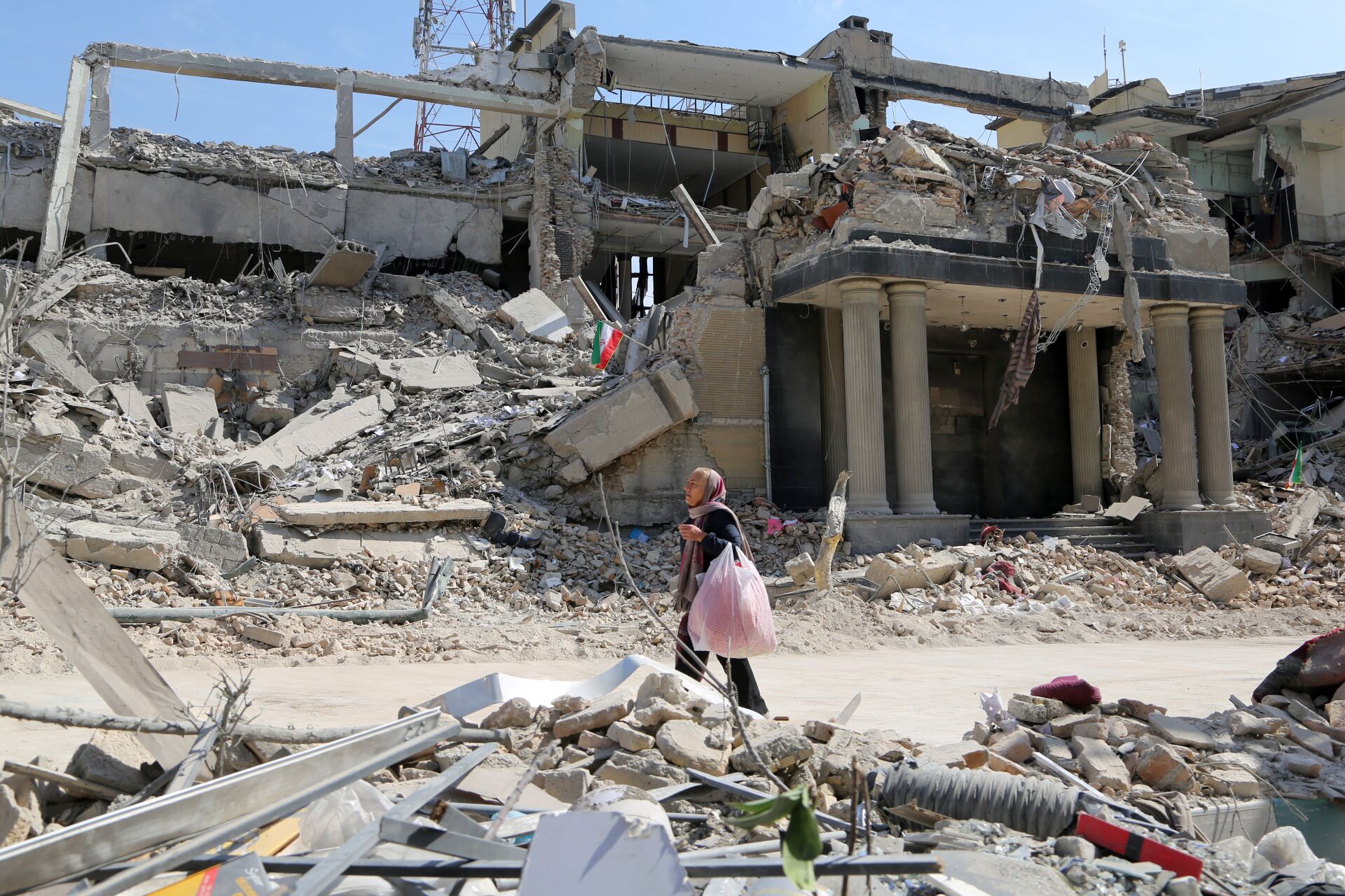 A person walks across rubble in front of a severely damaged concrete building, carrying bags amid debris and twisted metal in an urban setting.